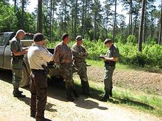 men in uniform standing around a truck on the side of a dirt road with trees behind them