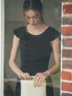 a woman in black shirt holding a white purse near brick wall and window sill