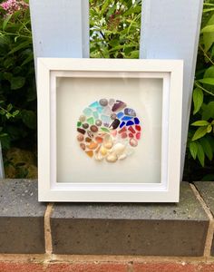 a white frame holding a multicolored glass mosaic in front of some bushes and flowers