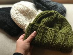 a hand is pointing at three knitted hats on a couch with one being held up to the camera