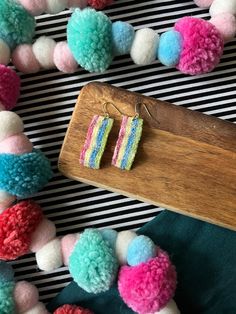 colorful pom - pom earrings are displayed on a wooden board with other pompoms