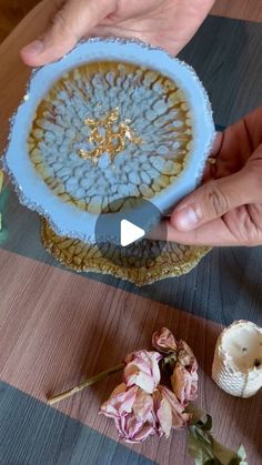 two hands are holding a blue and white plate with flowers on the table next to it