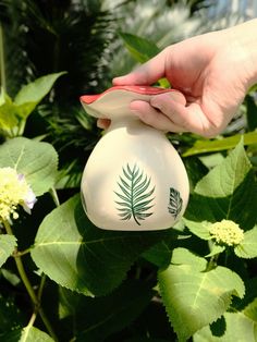 a hand holding a white vase with a green leaf on it in front of some flowers