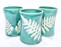 three green cups with white leaves on them sitting next to each other in front of a white background