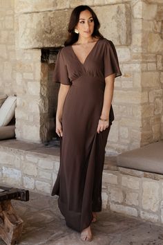 a woman standing in front of a stone fireplace wearing a brown dress with short sleeves