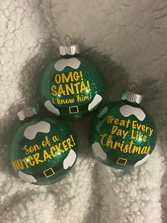 three green ornaments with words on them sitting on a white blanket in the shape of christmas balls