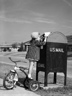 a woman in a plaid dress standing next to a mailbox with a bicycle parked by it