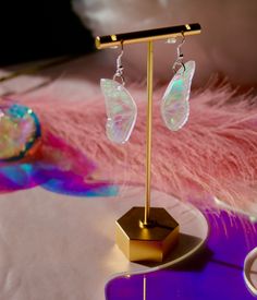 a pair of butterfly shaped earrings on a gold plated stand with pink feathers in the background