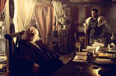 an old man sitting in a chair next to a table filled with books and other items