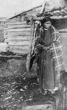 an old black and white photo of a woman standing in front of a log cabin