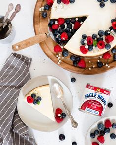 a cheesecake topped with berries and blueberries on a wooden platter next to a bowl of yogurt