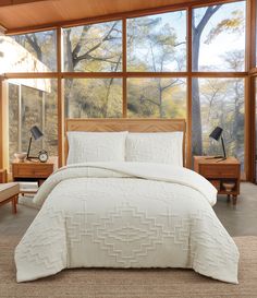 a bed with white comforter and pillows in front of large windows overlooking the woods