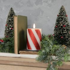 a red and white candle sitting on top of a table next to a book near christmas trees