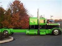 a large green semi truck parked in a parking lot next to some trees and bushes