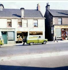 an old photo of people walking down the street in front of some shops and cars