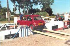 an old red car is parked in front of a white fence with people standing on it