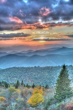 the sun is setting over mountains and trees in the foreground, with colorful foliage below