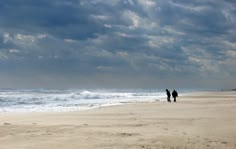 two people are walking along the beach under cloudy skies