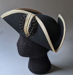 "This military style three cornered tricorn hat is made from top quality wool felt. The edges have been trimmed even and the sides held up or \"cocked\". It has been trimmed around the brim with gold fold-over braid. It is also available with black or white braid. Plain untrimmed hats are listed separately. Gold tone of edging may vary due to availability. Attached is a 3.5 \" black and gold, pleated double cockade with gold fleur de lis button below. Other buttons are available in the variation Formal Black Brimmed Costume Hats And Headpieces, Formal Black Costume Hats With Curved Brim, Black Formal Costume Hat With Curved Brim, Formal Black Brimmed Costume Hat, Vintage Black Hat For Costume Party, Black High Crown Hat For Formal Occasions, Black High Crown Costume Hat For Formal Occasions, Formal Black High Crown Costume Hat, Classic Black Hat For Costume