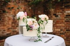 three white wedding cakes with flowers on top sitting on a table next to a brick wall