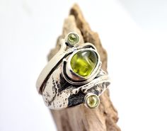 Lovely raw tumbled peridot gemstone and two small round peridots on a freeform leaf band handcrafted from sterling silver The band of the ring it is formed as a leaf and is wrapping the finger ending to small helixes. It is one of a kind ring. The shank of the ring is unusual, textured and have a light oxidation. It is designed on Lesbos island by us Nikiforos and Nelly inspired from walks into the greek nature. The ring is one of a kind and is made in US size 6 ,European 52 and UK M round perid Nature-inspired Natural Stones Jewelry For Anniversary, Unique Peridot Jewelry For May Birthstone, Peridot Rings With Natural Stones For Gifts, Handmade Peridot Jewelry For May Birthstone, Handmade Peridot Rings As Gift, Handmade Peridot Rings For Gift, Adjustable Peridot Jewelry For Anniversary, Peridot Birthstone Ring Jewelry, Artisan Natural Stone Ring