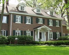 a large brick house with green shutters and white trim on the front door, surrounded by lush greenery