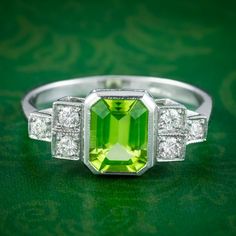 "A striking Art Deco inspired ring fashioned in platinum and bezel set with an emerald cut peridot in the centre weighing approx. 1.5ct. It has a bright green hue that is complemented perfectly by six twinkling brilliant cut diamonds arranged on stepped shoulder accents edged with milgrain. Peridot is the birthstone of August. They're considered a stone of summertime and were named \"the gem of the sun\" by the ancient Egyptians. Its natural lime-green colouring and inner light evoke the beauty Art Deco Green Ring, Gia Certified, Art Deco Green Ring Gia Certified, Platinum Green Diamond Ring, Green Diamond Platinum Ring Fine Jewelry, Fine Jewelry Green Diamond Platinum Ring, Green Octagon Diamond Ring, Fine Jewelry Green Diamond Ring In Platinum, Green Diamond Platinum Ring With Prong Setting, Green Platinum Diamond Ring