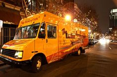 a yellow food truck parked on the side of a street at night with buildings in the background