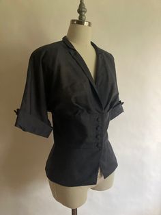 "1940s lightweight short sleeve jacket/blouse with pleated bust, covered buttons and lightly padded shoulders. Although the jacket appears to be black in the pictures, it is more of a slate grayMade by Wilshire of Boston, in excellent condition. The fabric is a bit of a mystery, it is lightweight with a striated texture, it could possibly be cotton or a form of rayon.  Shoulders 17\" across Chest 42\" (21\" across, doubled) Waist 34\" Hem 42\" Sleeve from neck to rolled cuff16\" Length from back of neck to hem 23\"" Tailored Short Sleeve Formal Tops, Elegant Fitted Single Breasted Top, Elegant Fitted Single-breasted Top, Tailored Short Sleeve Tops For Office, Fitted Blouse With Lapel Collar And Buttons, Classic Tailored Tops For Evening, Fitted Blouse With Buttons And Lapel Collar, Classic Fitted Blouse With Lapel Collar, Fitted Collared Blouse With Hidden Button Closure