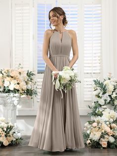 a woman standing in front of flowers wearing a long gray bridesmaid dress and holding a bouquet