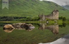 a castle sitting on top of a lush green hillside next to a lake in front of a mountain