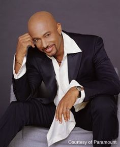 a balding man in a black suit and white shirt sitting on a gray chair