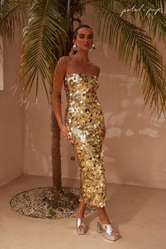 a woman standing next to a palm tree wearing a gold sequin dress with silver heels