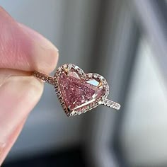 a pink diamond heart shaped ring being held up by someone's hand in front of a window