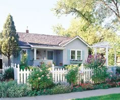 a white picket fence in front of a blue house with trees and flowers around it