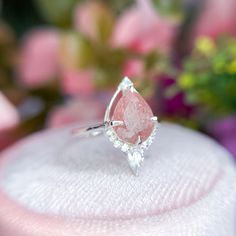 a pink diamond ring sitting on top of a white cloth covered table with flowers in the background