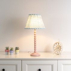 a lamp sitting on top of a white dresser next to a clock and potted plants