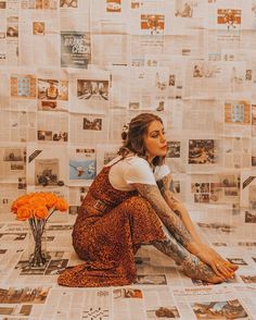 a woman sitting on the ground in front of a wall covered with newspapers and orange flowers