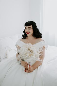 a woman sitting on top of a white bed holding a bouquet in her right hand