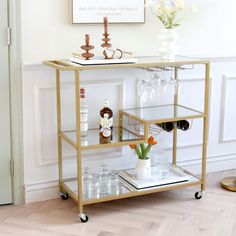 a gold bar cart with wine glasses and bottles on it in front of a white wall