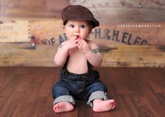 a baby sitting on the floor wearing a hat and holding his hands to his mouth