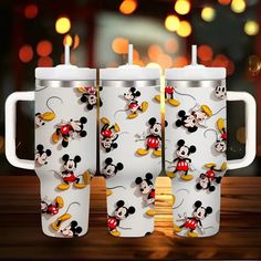 three mickey mouse travel mugs sitting on top of a wooden table with lights in the background