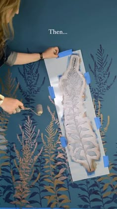 a woman is painting an image on the wall with blue and gold leaves in front of her