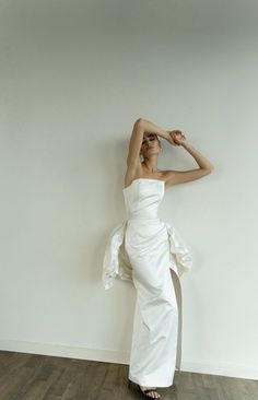 a woman in a white dress leaning against a wall with her hands on her head