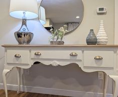 a white table with two vases on it and a mirror over the top that has flowers in front of it