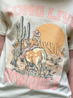 Cream tshirt with western print of cowgirl on horse with writting " long live cowgirls" Western-style Letter Print Tops For Western-themed Events, Western Style Screen Print Tops For Western-themed Events, Western Tops With Screen Print For Western-themed Events, Western Style Tops With Screen Print For Western-themed Events, Western Style T-shirt For Rodeo, Long Live Cowgirls Shirt, Western Style Screen Print T-shirt For Western-themed Events, Country Style Graphic Print Top For Rodeo, Western Rodeo Tops With Letter Print