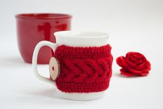 a knitted coffee cup cozying next to a red mug coziet with a rose on the side