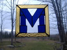 a blue and gold stained glass letter m hanging from a tree in front of a grassy area