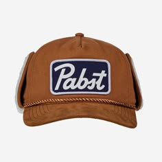 Stay warm in style with the Pabst Blue Ribbon Beer Shearling Hat with Ear Flaps in Tan. The classic Pabst logo adds a fun touch for fans of the iconic brand. This cozy hat features soft shearling lining and ear flaps to keep you snug on chilly days. Perfect for winter outings or casual wear, this hat is both practical and stylish.