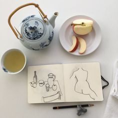 an open notebook next to a tea pot and plate with apples on it, along with a drawing of a woman's torso